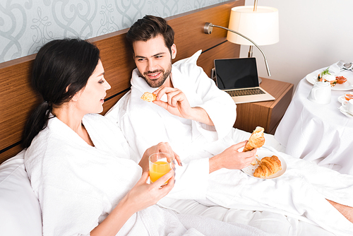 cheerful man holding croissant while looking at attractive woman with glass of orange juice