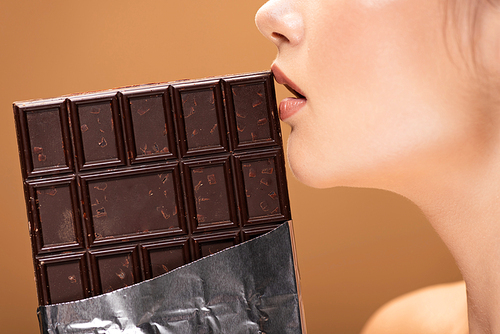 partial view of beautiful young woman holding chocolate bar in foil near lips isolated on beige
