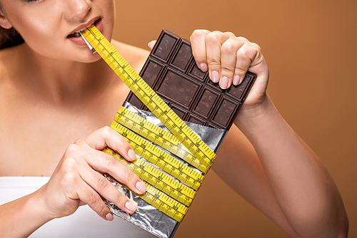 partial view of young woman holding yellow measuring tape in mouth and chocolate bar isolated on beige