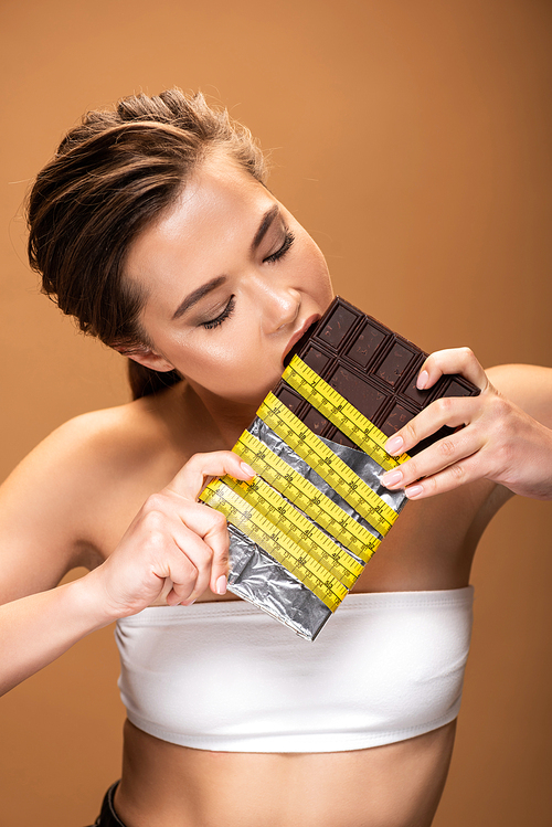 young woman with yellow measuring tape eating chocolate isolated on beige
