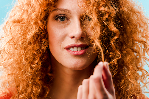 portrait of smiling woman holding red curly hair isolated on blue