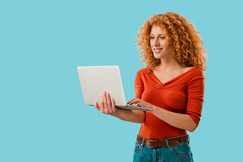 smiling redhead woman using laptop isolated on blue