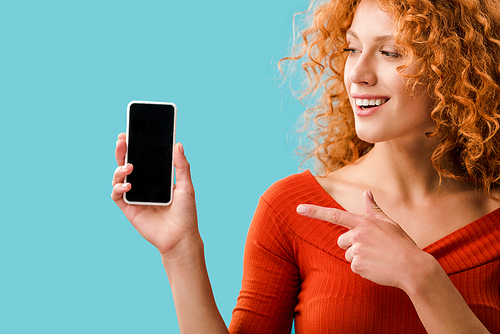 smiling redhead girl pointing at smartphone with blank screen isolated on blue