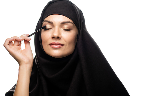 young Muslim woman in hijab applying mascara on eyelashes with closed eyes isolated on white