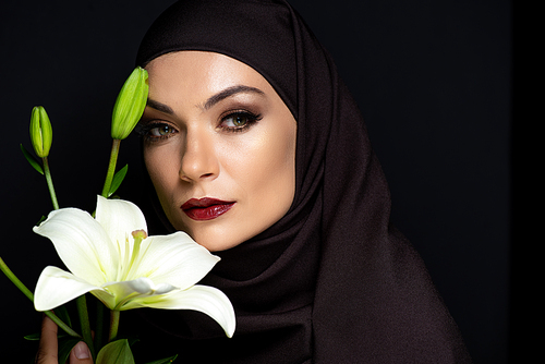 beautiful Muslim woman in hijab with smoky eyes and red lips holding lily isolated on black