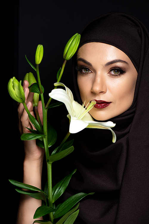 attractive Muslim woman in hijab with red lips holding lily isolated on black