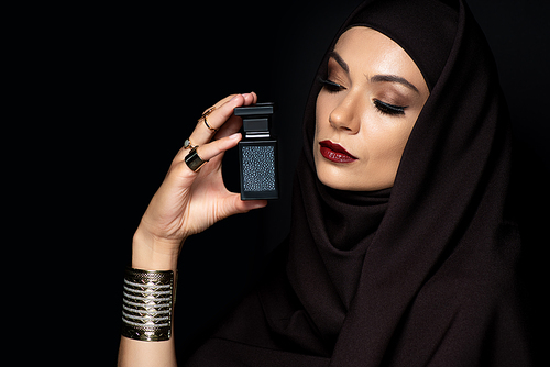 beautiful Muslim woman in hijab with makeup in golden jewelry holding perfume isolated on black