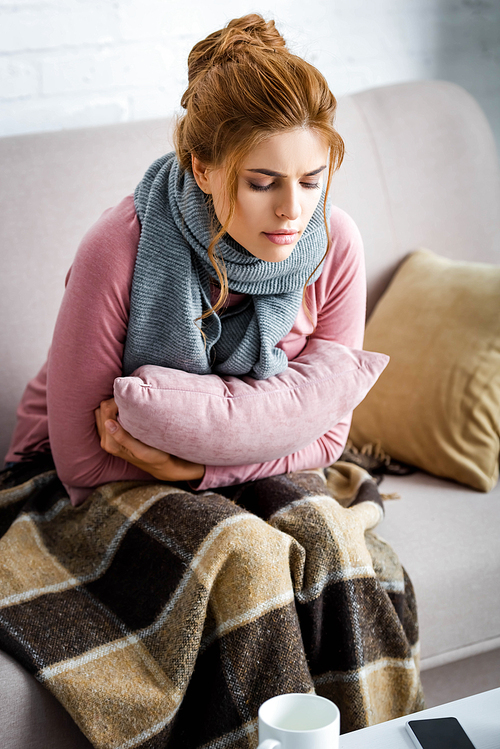 attractive and ill woman with grey scarf sitting on sofa and holding pillow