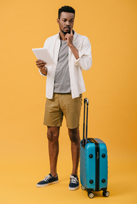 pensive african american man holding digital tablet and standing near luggage on orange