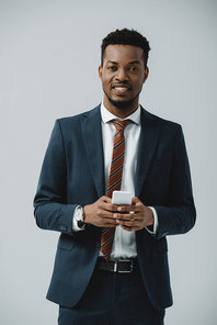 positive african american man in suit using smartphone isolated on grey