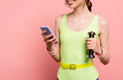 cropped view of cheerful sportswoman holding dumbbell and using smartphone on pink