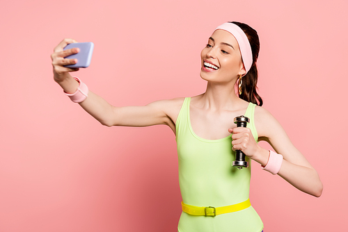 cheerful sportswoman holding dumbbell and taking selfie on pink