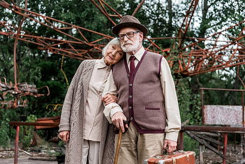 senior man in hat holding suitcase while standing with wife near abandoned carousel