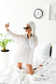 stylish woman in shirt, sunglasses, jewelry and with towel on head taking selfie in bed