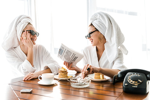 stylish happy women in bathrobes, sunglasses and jewelry with towels on heads smoking cigarette, laughing and reading newspaper at morning