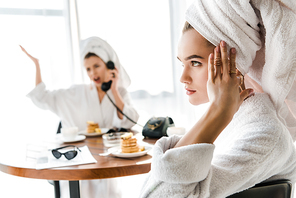 selective focus of exhausted stylish woman in bathrobe and jewelry with towel on head touching head while emotional friend talking on phone