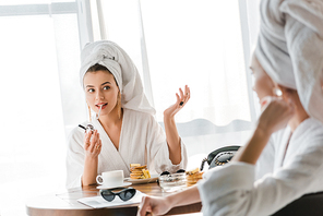 selective focus of smiling stylish woman in bathrobe and jewelry with towel on head lighting up cigarette while looking at friend