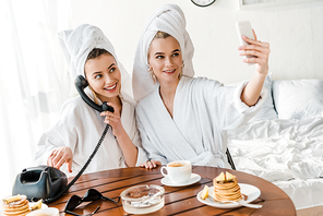 stylish women in bathrobes and jewelry with towels on heads talking on retro phone and smiling while taking selfie