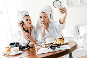 selective focus of stylish women in bathrobes and jewelry with towels on heads talking on retro phone and grimacing while taking selfie