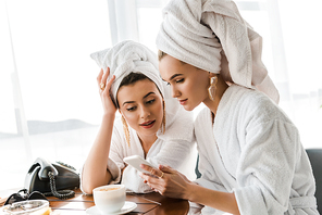 stylish women in bathrobes and jewelry with towels on heads using smartphone