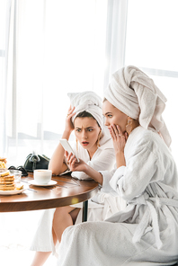 stylish surprised women in bathrobes and jewelry with towels on heads using smartphone during breakfast