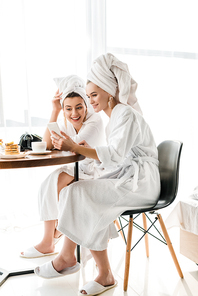 stylish smiling women in bathrobes and jewelry with towels on heads using smartphone during breakfast