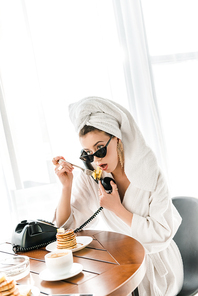 stylish woman in bathrobe, sunglasses and jewelry with towel on head talking on vintage telephone while eating pancakes