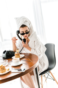 stylish woman in bathrobe, sunglasses and jewelry with towel on head talking on retro telephone while eating pancakes