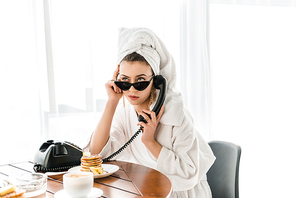 stylish woman in bathrobe, sunglasses and jewelry with towel on head talking on retro telephone while having breakfast