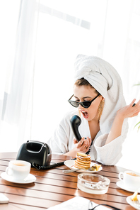 irritated stylish woman in bathrobe, sunglasses and jewelry with towel on head screaming while talking on retro telephone