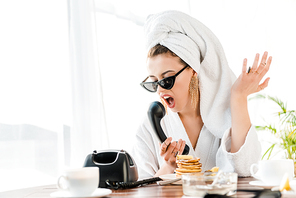 irritated stylish woman in bathrobe, sunglasses and jewelry with towel on head screaming and gesturing while talking on retro telephone