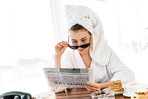 stylish woman in bathrobe, sunglasses and jewelry with towel on head reading newspaper at morning