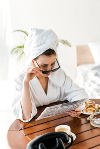 woman in bathrobe, sunglasses and jewelry with towel on head reading newspaper at morning