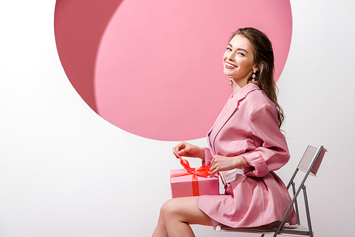 happy woman sitting on chair and holding present on white and pink