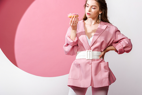 attractive woman standing with hand on hip and looking at sweet doughnut on white and pink