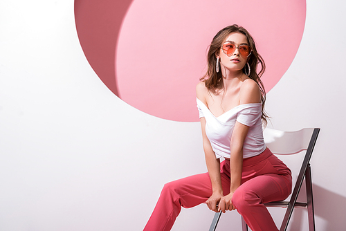 attractive woman in sunglasses sitting on chair on white and pink