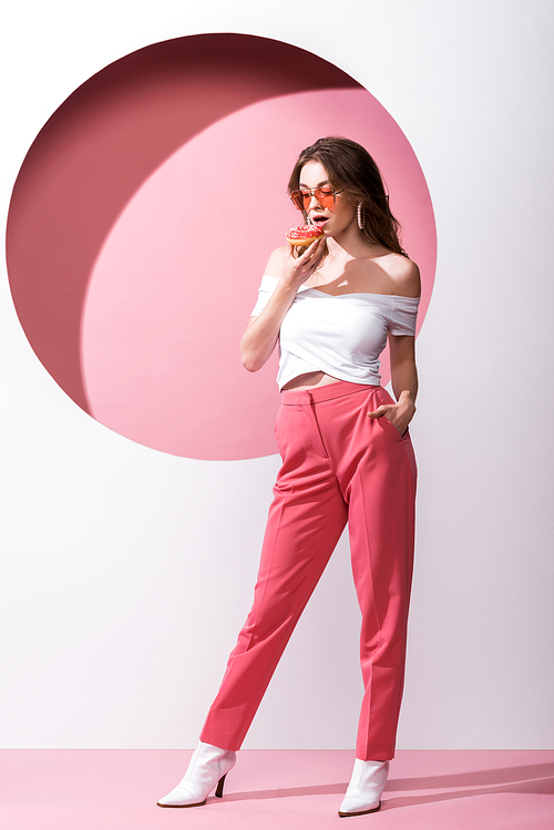 beautiful girl holding tasty doughnut and standing with hand in pocket on pink and white