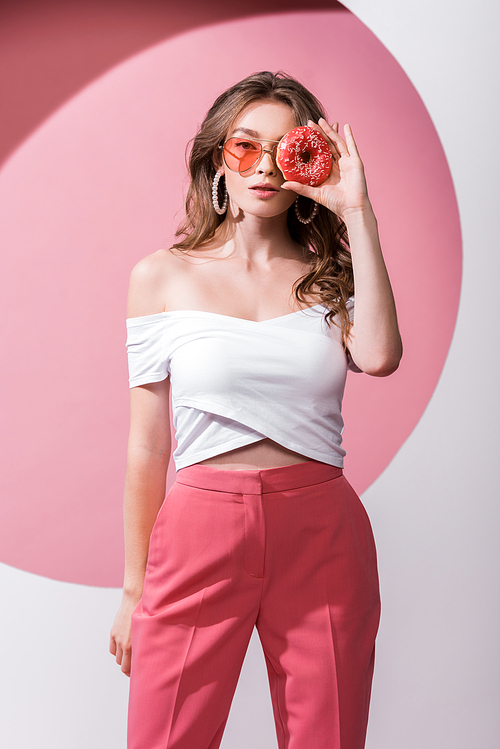 beautiful girl covering face with tasty doughnut and standing on pink and white