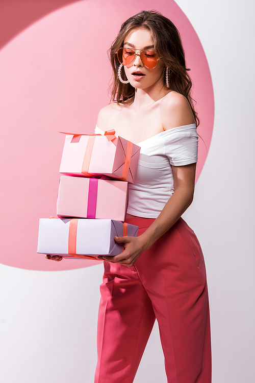 surprised woman in sunglasses holding presents on pink and white