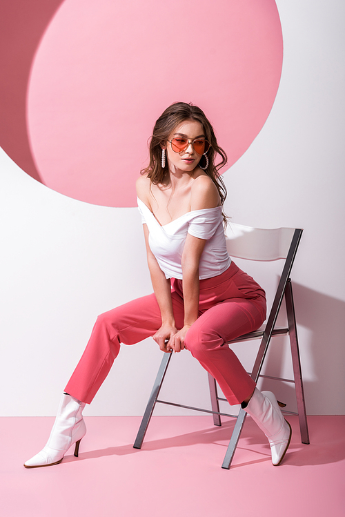 attractive woman in sunglasses sitting on chair on pink and white