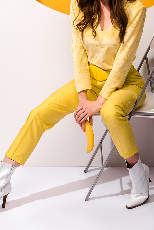 cropped view  of woman sitting on chair and holding banana on white and orange