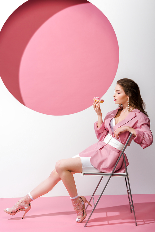 attractive woman sitting on chair and holding sweet doughnut on pink and white