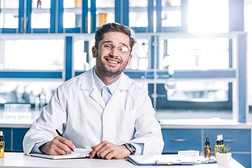 happy man in glasses and white coat sitting at desk in clinic