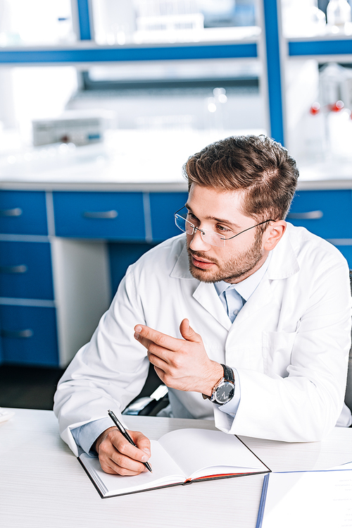 handsome doctor in glasses writing in notebook and gesturing in clinic