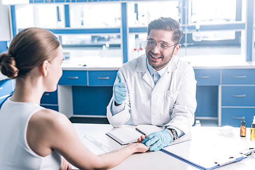 selective focus of happy doctor showing thumb up and holding hand of woman in clinic