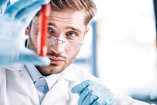 selective focus of handsome immunologist in glasses holding test tube with red liquid in clinic