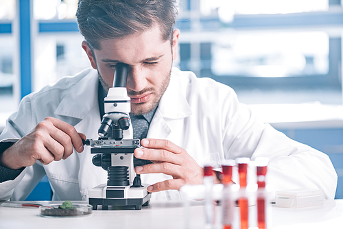 selective focus of bearded biochemist looking through microscope in laboratory