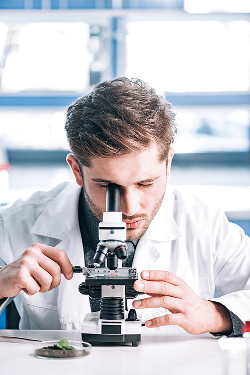 selective focus of bearded biochemist in white coat looking through microscope in laboratory