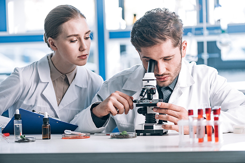 selective focus of biochemist looking through microscope near attractive coworker