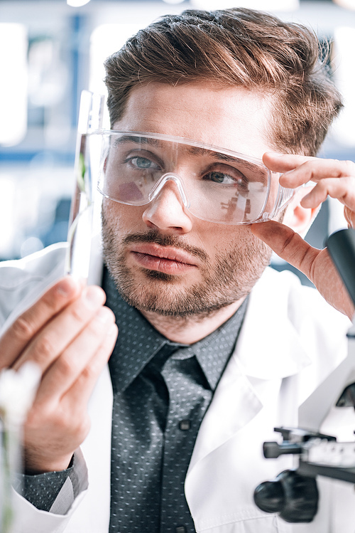 selective focus of handsome biochemist touching goggles and looking a test tube